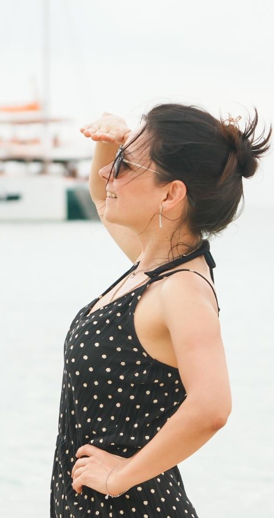 Goreti on a beach in profile, hand shading her eyes, looking out to sea with a boat on the water behind her.