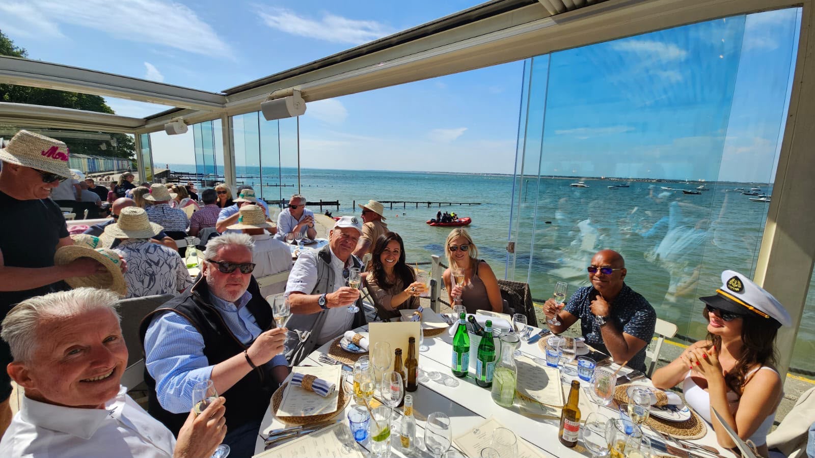 A group raising glasses at a long table by the sea on a bright day.