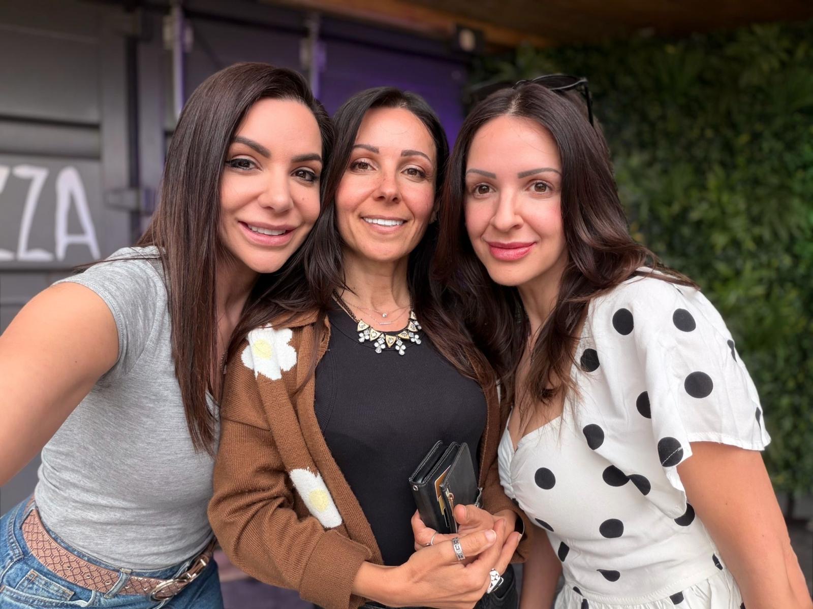 Goreti standing close with two other women outdoors, all three smiling at the camera.